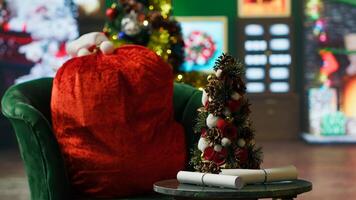 Close up of small Christmas tree on table next to red Santa bag full of presents. Focus on letter scrolls received by Santa Claus from kids in workshop adorn with holiday props, handheld camera shot photo