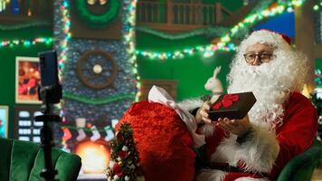 Man playing Santa Claus filming with mobile phone on tripod, taking gifts from red bad to show children in Christmas set backdrop with stockings, fireplace, ornaments and other holiday props, camera A photo