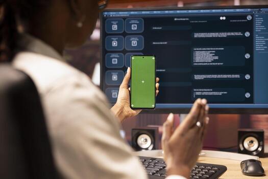 Woman seated at desk using AI LLM chatbot software and green screen phone in modern urban home office. Person uses large language model program on computer and mockup smartphone in apartment photo