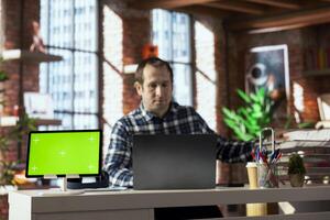 Isolated screen tablet in front of worker at home office desk picking paper file from stack, using laptop, drafting contracts. Close up of mockup electronic device next to employee checking paperwork photo