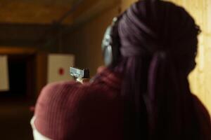 Woman in firing range doing shooting activity with pistol, wearing earmuffs to protect hearing. Person in shooting gallery training with firearm, using safety gear to prevent damage from gunshots photo
