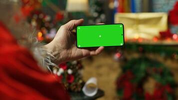 Person dressed as Santa Claus reading letters from kids on chroma key smartphone. Senior man portraying Christmas character answering children using isolated screen mobile phone, camera A close up photo
