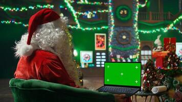 Santa Claus performer typing on green screen laptop, preparing shipping parcel with gift to be sent to kids. Man uses mockup notebook, scheduling goods delivery, preparing to send presents to children photo
