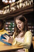 Surprised teenage girl enjoying mandatory literature book in the library, feeling amazed by the lecture story. Schoolgirl reading literary work from the annual school curriculum, academic success. photo