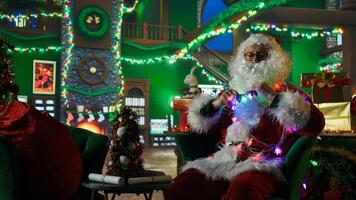 Santa Claus entangled in led lights while adorning workshop for Christmas, preparing it for families visiting. Costumed man knotted in xmas lights while doing holiday season decorations, camera A photo