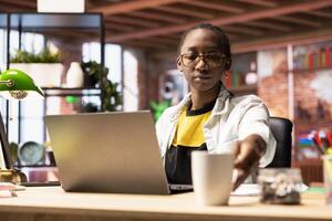 Woman in home office drinking coffee in the morning and browsing internet, researching information. African american teleworker starting job shift with hot beverage, preparing to do work tasks photo
