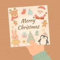Hand holding cute pastel Merry Christmas card with Santa, bunny, penguin, reindeer, gingerbread cookie and Christmas tree on wooden table background in kawaii cartoon style vector
