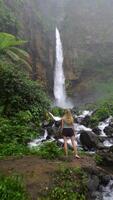 Blonde woman standing in front of the majestic Kapas Biru waterfall in East Java, Indonesia, surrounded by lush jungle, mossy rocks, and flowing river. video