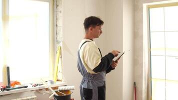Professional builder wearing overalls using digital tablet during renovation works in apartment, checking project plan or ordering building materials video