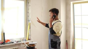 Construction worker wearing overalls is making a phone call and gesturing with his hand while standing in a room undergoing renovation, with tools and materials visible on the windowsill video