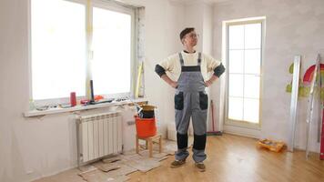 Painter in overalls stands proudly in a freshly renovated room, hands on hips, surrounded by a paint bucket and various tools, showcasing a sense of accomplishment and creativity video