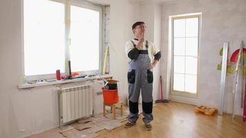 Painter in overalls thoughtfully evaluating next steps in a renovated room, surrounded by painting tools and partially completed work, illustrating home improvement progress video