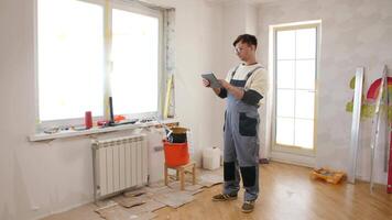 Painter in overalls and glasses using a digital tablet in a room under renovation, surrounded by painting tools and materials for a home improvement project video