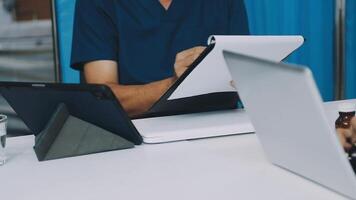 A man in scrubs is sitting at a table with a laptop and tablet video