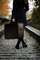 Woman in Black coat with Vintage suitcase walks along railway path in autumn, surrounded by vibrant fall leaves, capturing essence of travel and exploration. photo