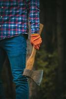 Cropped view of man Lumberjack in plaid shirt and jeans holds axe, ready for work in forest. photo