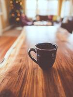 A dark mug of coffee sits on a rustic wood table in a cozy living room with a blurred Christmas tree in the background. photo