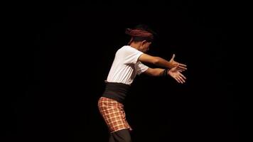 A male dancer performing on stage, dressed in traditional attire with a headband and a patterned sarong. The background is dark, highlighting the dancer's movements and posture. video