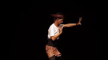 A male dancer performing a traditional dance on stage, wearing a white shirt and patterned sarong. The background is dark, highlighting the dancer's movements and expressions. video