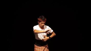 A male dancer performing on stage with a dramatic pose, wearing traditional attire and a headband. The background is dark, highlighting the dancer's movements and expression. video