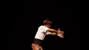 A young dancer performing on stage, showcasing traditional dance moves. The dancer wears a white shirt, a patterned sarong, and a headband, with a dramatic pose against a dark background. video