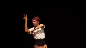 A male dancer performs on stage, wearing traditional attire with a headband and a checkered sarong. The background is dark, emphasizing the dancer's movements and expressions. video