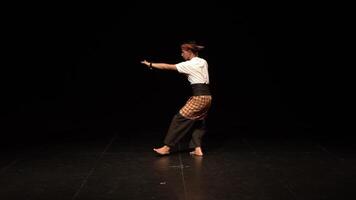 A solo dancer performing on stage, wearing traditional attire with a patterned sarong and a simple white shirt. The stage is dimly lit, focusing on the dancer's movements. video