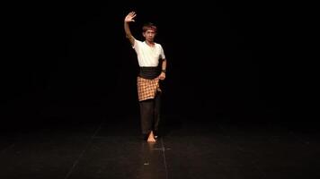 A dancer performing on stage, showcasing traditional movements in a dark setting. The dancer wears a white shirt and a patterned sarong, emphasizing cultural expression through dance. video