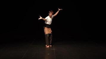 A male dancer performing a traditional dance on a dark stage, wearing a white shirt, black sash, and patterned sarong. The dancer is in a dynamic pose, showcasing movement and expression. video