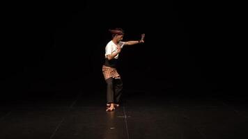 A male dancer performing on stage, dressed in traditional attire with a checkered sarong and a white shirt. The background is dark, highlighting the dancer's movements. video