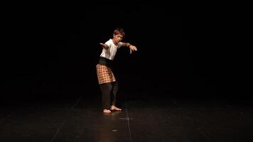 A dancer performing on a dark stage, showcasing traditional movements. The dancer wears a white shirt and a patterned sarong, with a focused expression, emphasizing cultural dance. video
