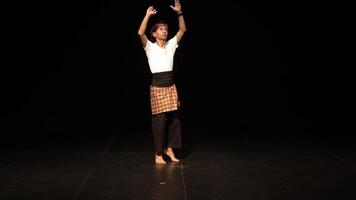 A male dancer performing on stage, wearing traditional attire with a checkered sarong and a white shirt. The background is dark, highlighting the dancer's movements and expressions. video