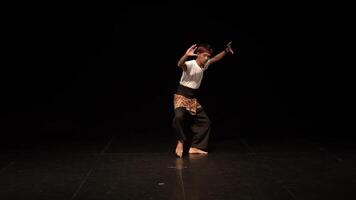 A male dancer performing on stage, dressed in traditional attire with a checkered sarong and a headband. The stage is dimly lit, focusing on the dancer's movements. video