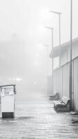 Monochrome Urban Fog A Quiet Streetscape Veiled in Dense Mist, Featuring Empty Benches, Glowing Streetlights, and a Newspaper Stand. photo