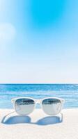 Stylish white sunglasses rest on pristine white sand, overlooking a tranquil blue ocean under a clear summer sky, embodying perfect tropical vacation relaxation. photo