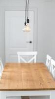Bright and inviting minimalist dining room with a rustic wooden table, classic white chairs, and modern Edison-style pendant lights illuminating the space. photo