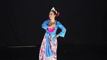 A dancer performing on stage, wearing a colorful traditional costume with floral patterns. The background is dark, highlighting the dancer's movements and attire. video