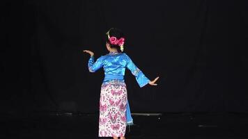 A dancer performing on stage, wearing a traditional blue top and floral patterned skirt. The background is dark, highlighting the dancer's movements and attire. video