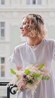 A young pretty girl is standing on the balcony smiling happily and looking around. A bouquet of flowers is in the girl's hand. Happy morning on the bedroom video