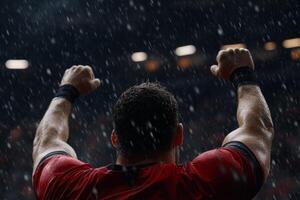 A muscular athlete raises arms in triumph amidst a rain shower during an intense night game. The stadium is alive with energy as fans cheer for the dramatic moment photo