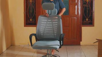 A person assembling a modern ergonomic office chair in a well-lit room with wooden doors in the background. The chair features a mesh design and adjustable components. video