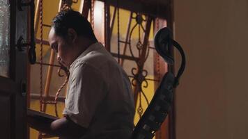A man reading a book while sitting in a chair near a window with decorative iron bars. The warm light from outside casts a soft glow, creating a cozy atmosphere. video