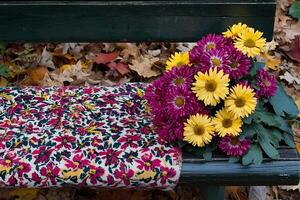 Autumnal Bouquet Resting on a Park Bench. A vibrant arrangement of purple and yellow chrysanthemums sits on a dark wooden bench amidst fallen leaves. photo