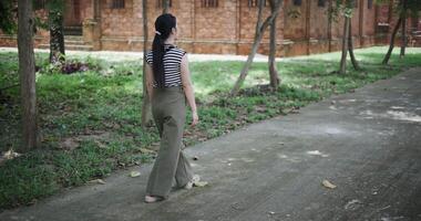 Handheld rear view, Young Asian woman in a striped shirt and green pants walks along a shady tree-lined path. Enjoy holiday travel. video