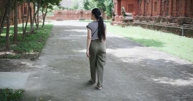Handheld rear view, Young Asian woman in a striped shirt and green pants walks along a shady tree-lined path video