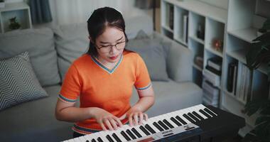 High angle view shot, Asian woman wearing glasses and an orange t-shirt is playing the keyboard with a smile in a beautifully decorated living room. video