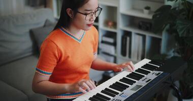 Handheld high angle view shot, Young Asian woman wearing glasses is playing the keyboard with a smile in a beautifully decorated living room. video