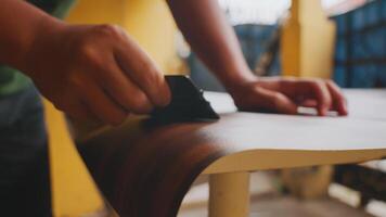 A close-up of a person's hand sanding a wooden surface, showcasing craftsmanship and attention to detail. The background features warm colors, suggesting a cozy workshop environment. video