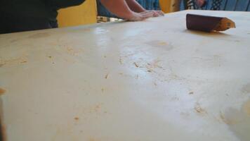 A close-up view of a wooden surface being sanded, showing scratches and dust particles. The background features blurred hands working on the surface, indicating a DIY or craft project. video
