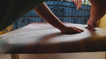 A close-up of a hand smoothing a wooden surface with a tool, showcasing craftsmanship and attention to detail. The background features a decorative blue gate and some hanging clothes. video
